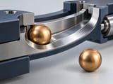 Conceptual CGI: warm bronze sphere on a smooth curved steel track, plus a separated intact sphere showing contact-material pairing rather than abstract hardness claims