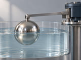 Conceptual CGI: A stainless hollow spherical float half immersed in a clean clear water vessel, simple metal lever fixed gently at top, visible realistic water meniscus and reflection, modest open level-sensing concept