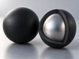 Conceptual CGI: Cutaway of a black rubber-coated sphere revealing one solid silver metal core with a uniform thick black elastomer shell, beside an intact matching sphere; coating bond shown simply, no exploded flakes