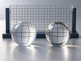 Conceptual CGI: Transparent acrylic sphere and transparent glass sphere beside a simple optical viewing grid, physically plausible different refractions; no labels or claimed measurement